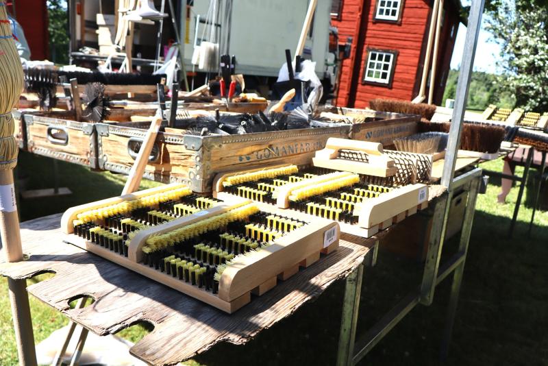 Ett urval av borstar och rengöringsredskap utställda på ett träbord utomhus på en marknad eller mässa.