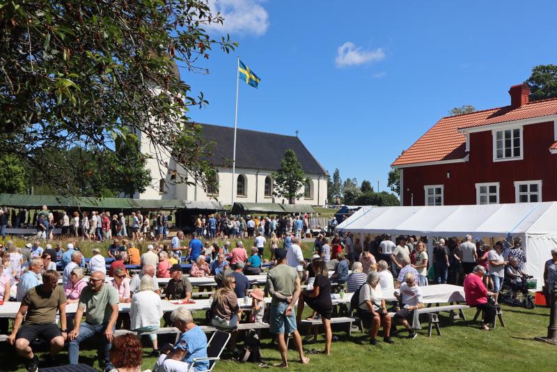 Människor samlas utomhus vid långbord för ett evenemang nära en vit kyrka, en svensk flagga och ett rött hus en solig dag.