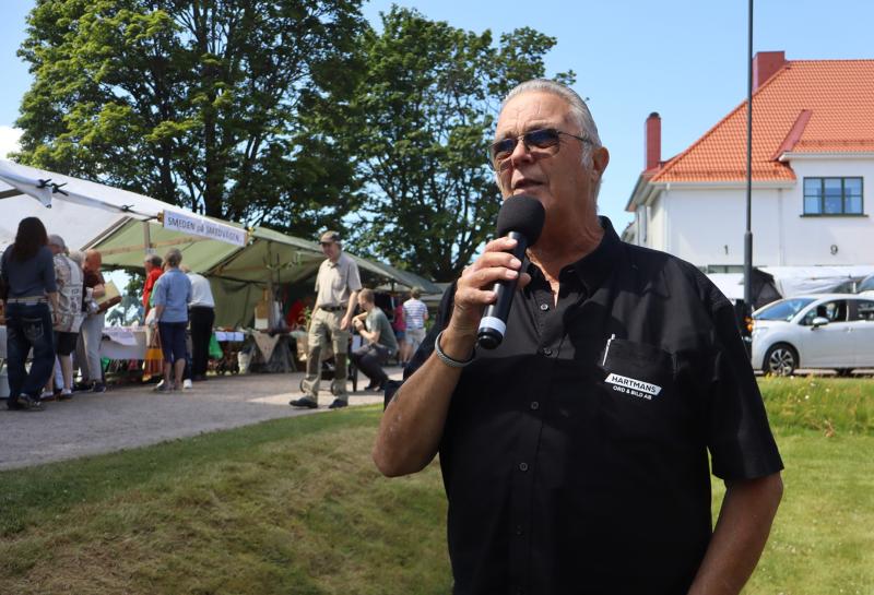 En man med solglasögon talar i en mikrofon vid ett utomhusevenemang, med tält, människor och en byggnad synliga i bakgrunden.