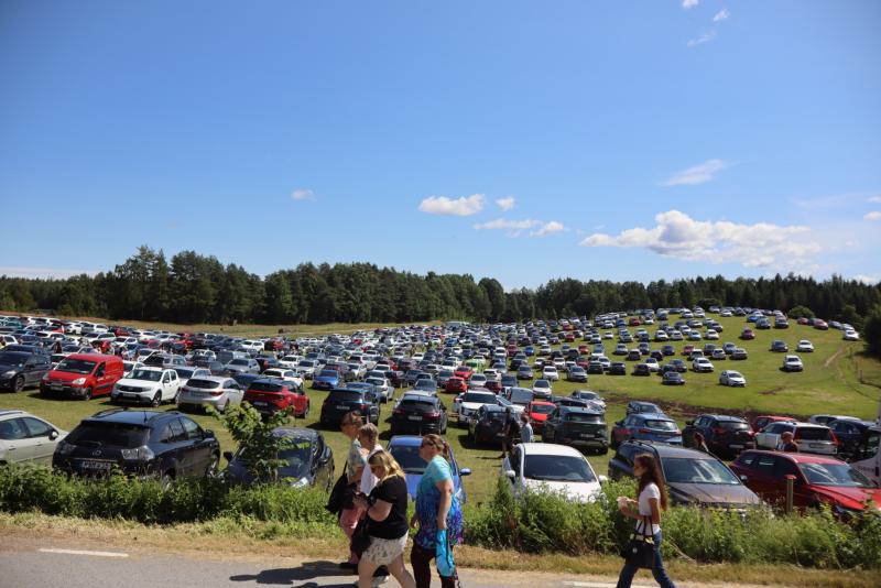 En stor gräsbevuxen äng som används som parkeringsplats, fylld med många parkerade bilar; flera personer går längs vägen i förgrunden under en klarblå himmel.
