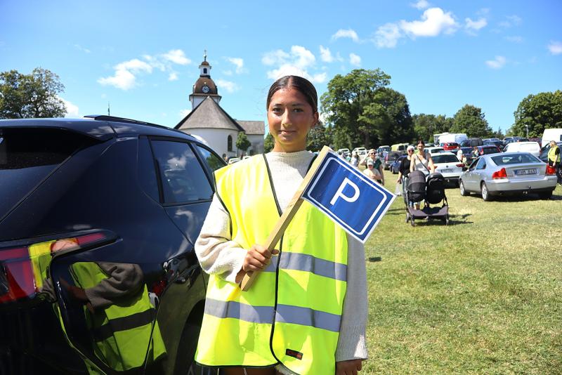En person i en gul säkerhetsväst som håller i en parkeringsskylt står bredvid parkerade bilar på en gräsbevuxen äng, med en kyrka och andra människor i bakgrunden.
