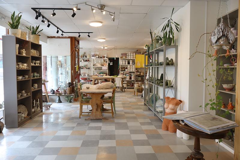 En keramikbutik med hyllor fyllda med keramik, krukväxter, ett träbord och naturligt ljus som strömmar in genom stora fönster.