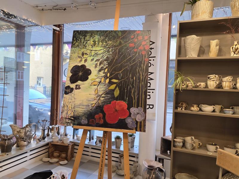 En tavla med stora blommor och bladverk på ett staffli är utställd i en butik där det finns keramik och porslin på hyllorna och där man från fönstret har utsikt över gatan utanför.