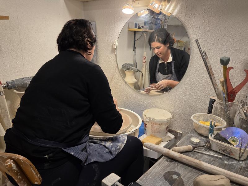 En person formar lera på en drejskiva i en ateljé, och personens spegelbild syns i en rund spegel ovanför arbetsbänken.