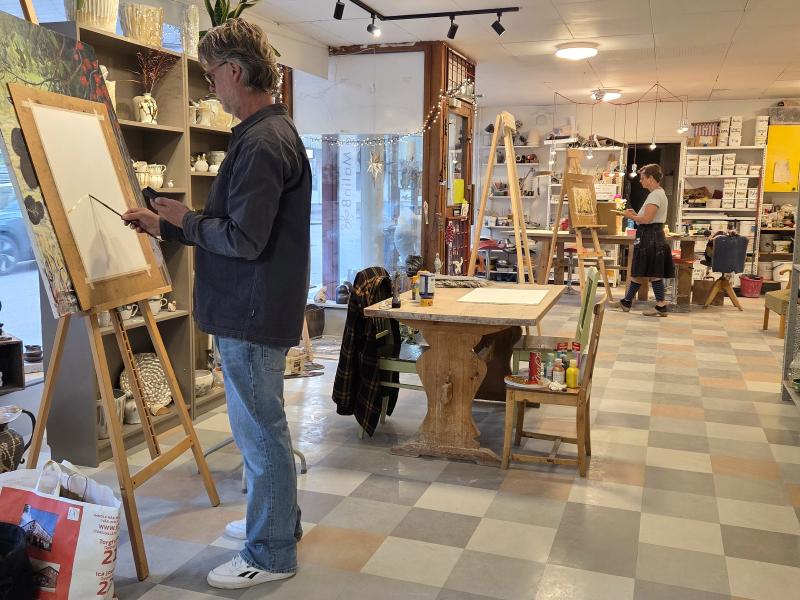 Två personer målar vid stafflier inne i en väl upplyst konstateljé och butik, där hyllor med keramik, konstmaterial och inredningsföremål syns runt om i rummet.