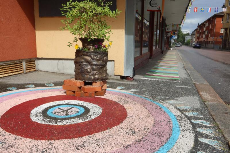 En målad trottoar med koncentriska cirklar och ett fredstecken, en blomlåda på tegelstenar och färgglada ränder som sträcker sig längs gatan i ett stadsområde.