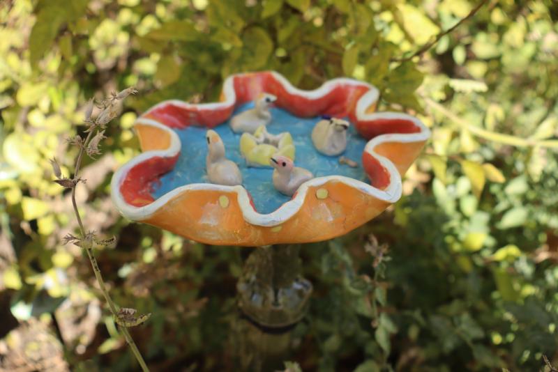 Ett färgglatt fågelbad med små ankafigurer flyter i blått vatten, omgivet av gröna, lövrika växter i en trädgård.