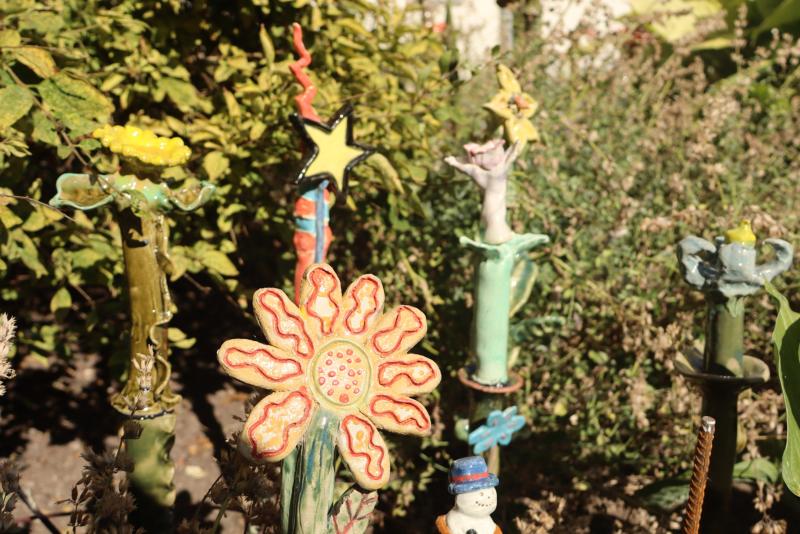 Färgglada keramikskulpturer i form av blommor och stjärnor står uppställda i en trädgård med grönt bladverk i bakgrunden.