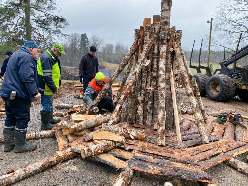 Flera personer i utomhuskläder monterar en stor träkonstruktion av stockar och plankor en molnig dag, omgivna av träd och utrustning.