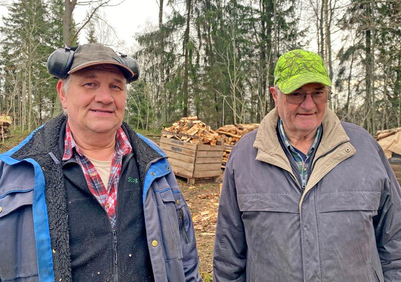 Två äldre män i arbetskläder står framför staplade vedlådor och träd, den ene med hörselkåpor och den andre med grön mössa.