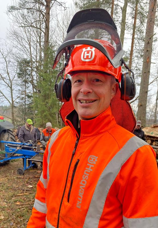 En man i orange Husqvarna-skyddskläder och hjälm står utomhus nära träd, medan andra arbetar i bakgrunden.