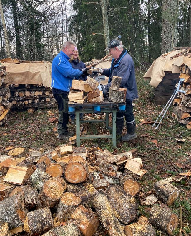Tre personer sågar vedträn med en såg vid en vedstapel utomhus, omgiven av staplar med avverkad ved och träd.