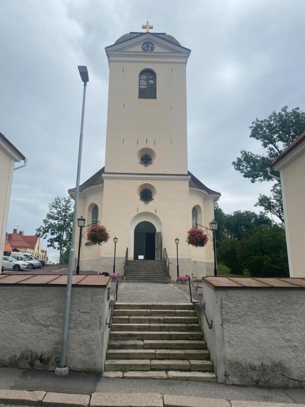 En gräddfärgad kyrka med ett högt entrétorn, välvd dörr, runda fönster och två hängande blomkorgar, som nås via en stentrappa.