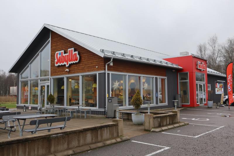 En Sibylla snabbmatsrestaurang med stora fönster, uteservering och synlig skyltning en molnig dag.