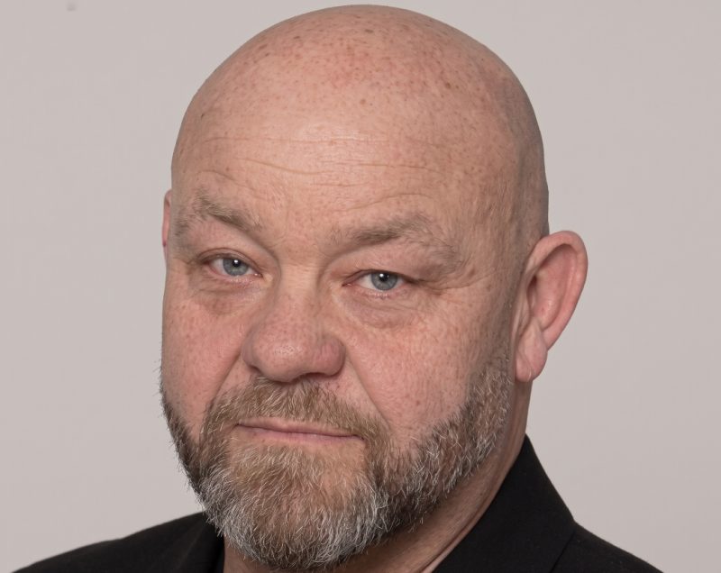 En skallig man i medelåldern med skägg, klädd i en mörk skjorta, som blickar rakt in i kameran med ett neutralt ansiktsuttryck mot en enfärgad bakgrund.