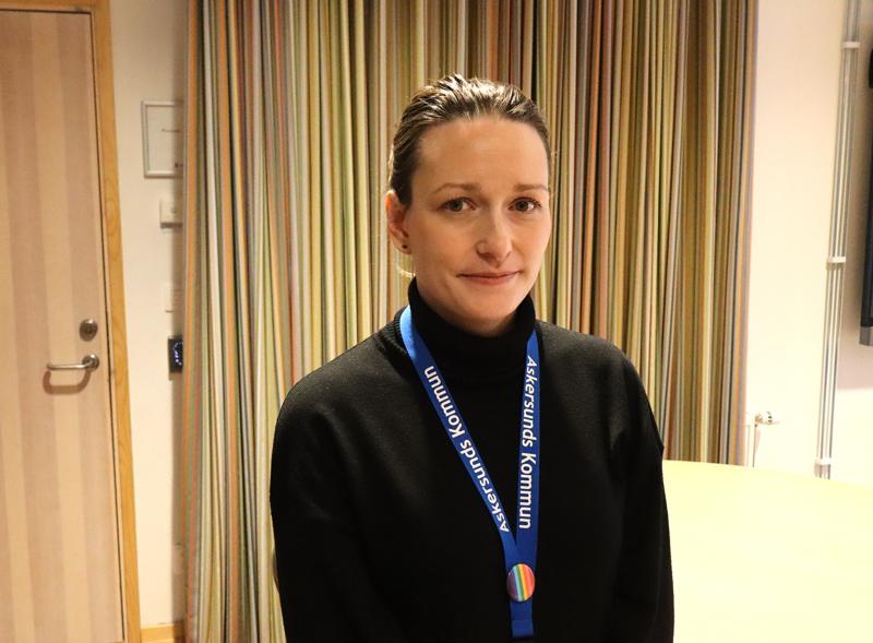 En kvinna med ett blått nyckelband står inomhus framför randiga gardiner, vänd mot kameran med ett neutralt uttryck.