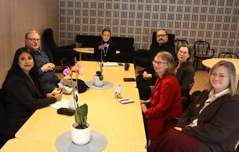 Sju personer sitter runt ett bord i ett mötesrum, med anteckningsböcker, muggar och orkidéer i kruka på bordet.