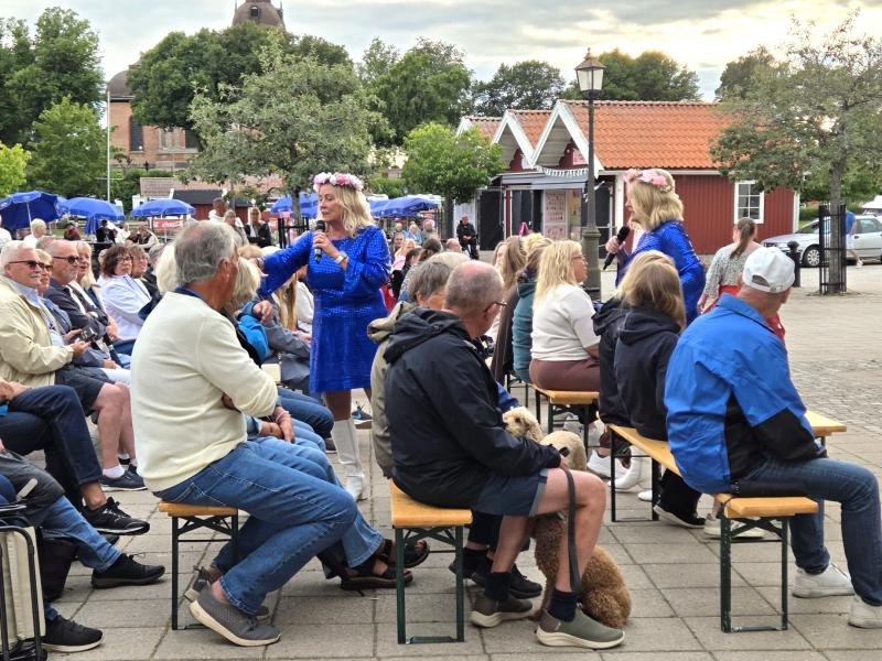 Två kvinnor i blå klänningar uppträder utomhus inför en sittande publik; vissa personer håller i hundar, och i bakgrunden syns träd, byggnader och marknadsstånd.