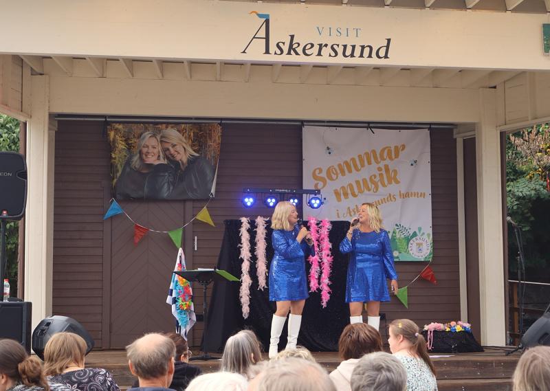 Två kvinnor i blå klänningar uppträder på en utomhusscen i Askersund, Sverige, med en banderoll med texten ”Sommar Musik” och en publik som sitter framför scenen.