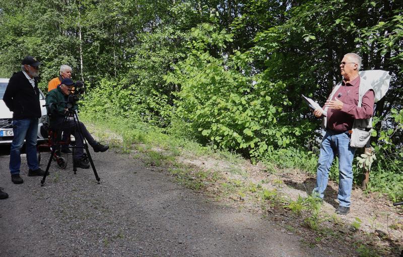 En man står på en grusgång och pratar med tre personer, varav en håller i en videokamera, med tät grön vegetation i bakgrunden.