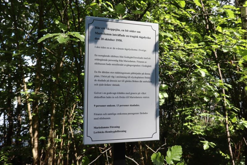 En informationsskylt i metall med text på svenska står bland gröna träd och lövverk i ett solbelyst skogsområde.