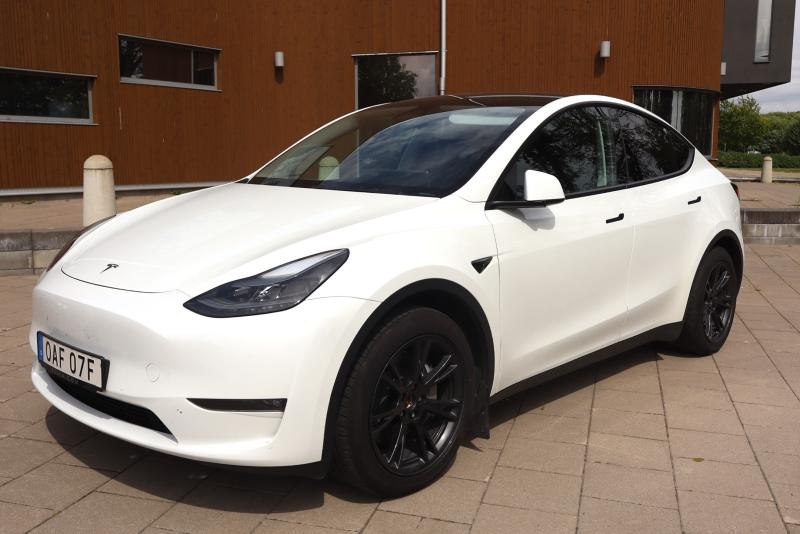 En vit Tesla Model Y står parkerad på en asfalterad yta framför en modern brun byggnad.
