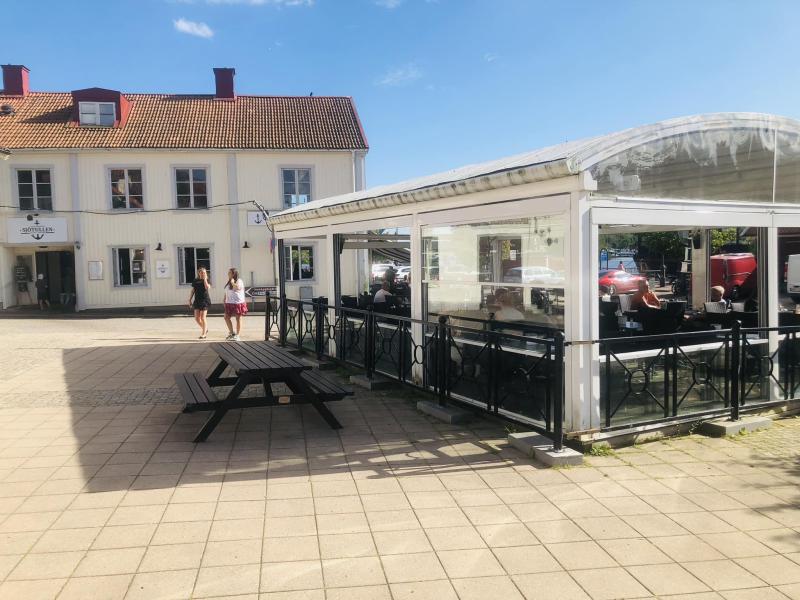 Två personer går mot en vit byggnad med rött tak och uteservering täckt av en klar plastkapell en solig dag. Ett picknickbord i trä står i förgrunden.