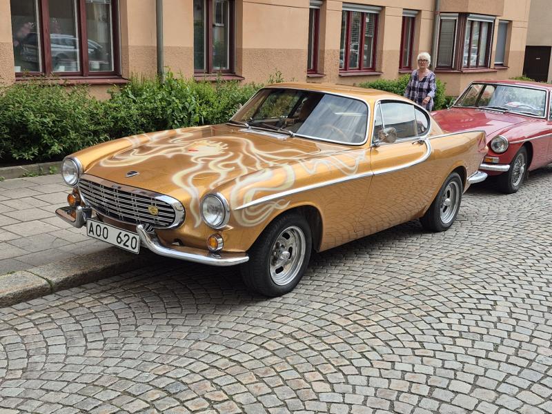 En guldfärgad klassisk bil med dekorativ färg parkerad på en kullerstensgata, med en annan veteranbil och en person i bakgrunden.