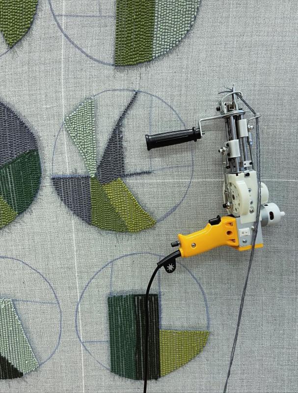 En handhållen tuftpistol används för att skapa geometriska, gröna och grå textilmönster i cirkulära konturer på ett grått tyg.