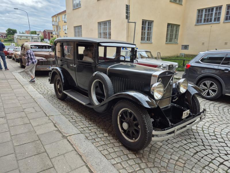 En svart veteranbil parkerad på en kullerstensgata med andra klassiska bilar och människor i närheten, mot bakgrund av beige byggnader.