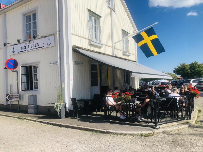 Folk sitter utanför en vit byggnad med svensk flagga och njuter av mat och dryck. En skylt lyder 