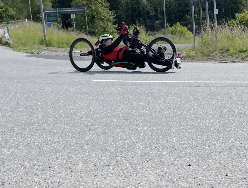 Person som cyklar på en liggande handcykel på en väg, bär en grön hjälm, solglasögon och en röd skjorta.