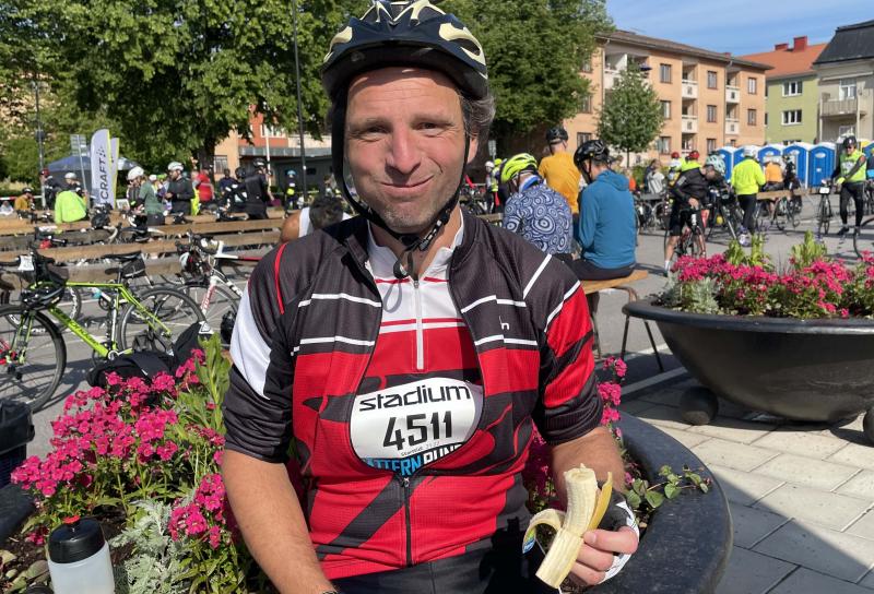 En cyklist som bär hjälm och numrerad tröja, märkt 4511, sitter på en bänk och äter en banan. Andra cyklister och cyklar syns i bakgrunden.