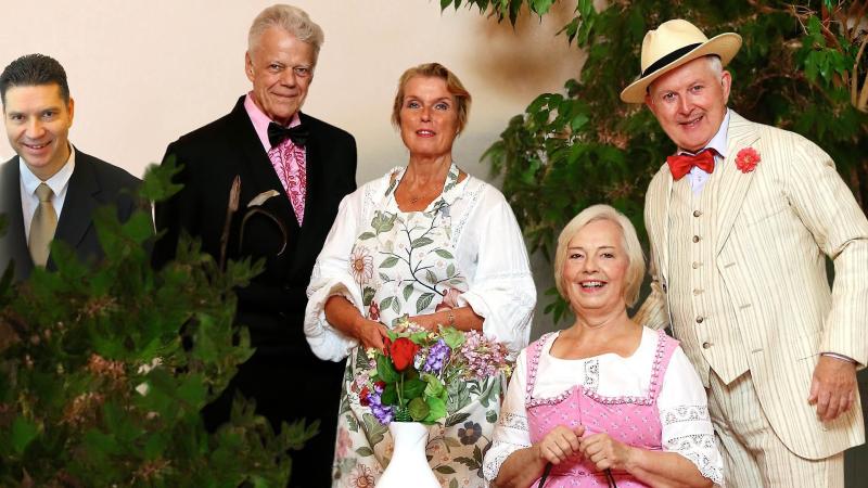 Fem vuxna, tre stående och två sittande, poserar inomhus. Tre är klädda i traditionell dräkt medan två bär kostym. Framför dem står en vas med blommor. Buskar och växter är synliga.