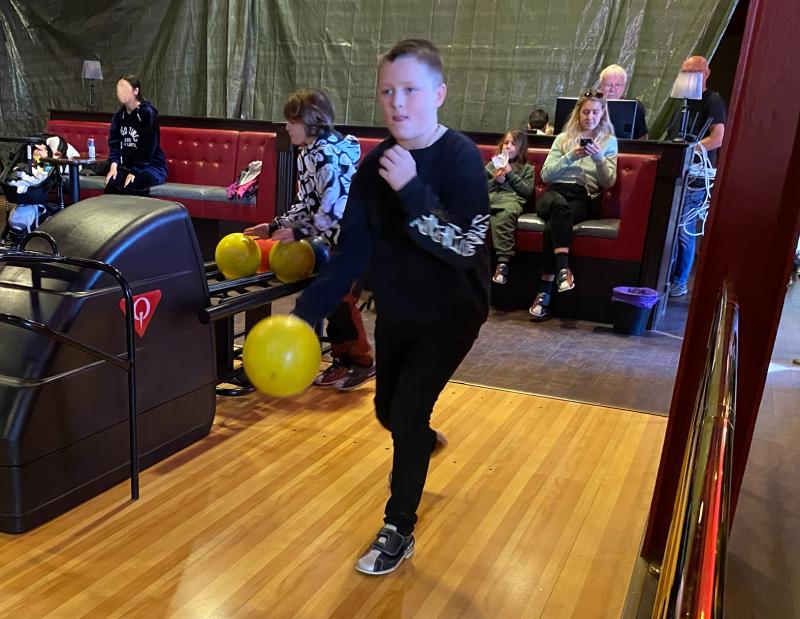 En pojke förbereder sig för att bowla med ett gult klot i en bowlinghall, medan flera personer sitter i bås i bakgrunden.