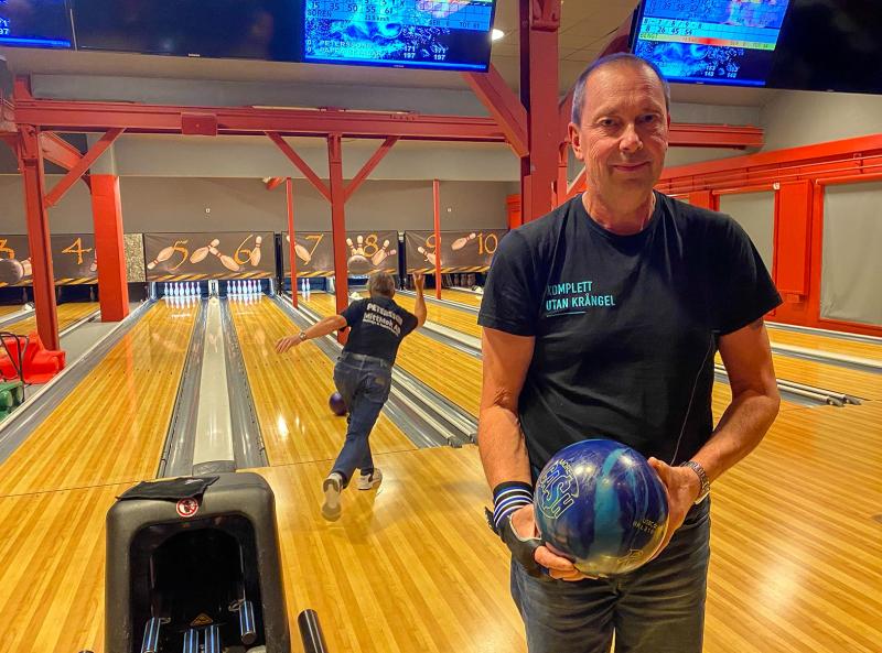 Två män befinner sig i en bowlinghall; den ene ska just släppa iväg ett bowlingklot längs banan, medan den andre står i förgrunden med ett blått bowlingklot i handen och vänd mot kameran.