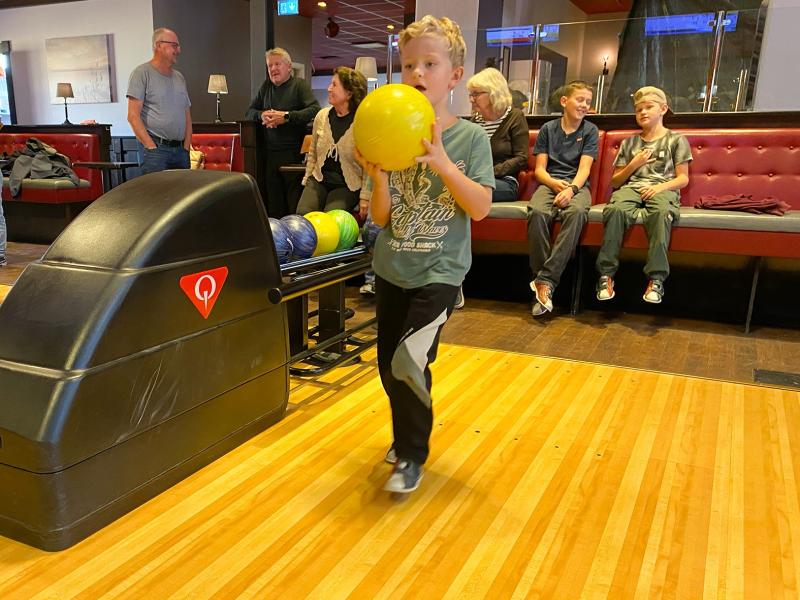 En pojke förbereder sig för att bowla med ett gult bowlingklot i en bowlinghall. Flera personer sitter och står i bakgrunden.