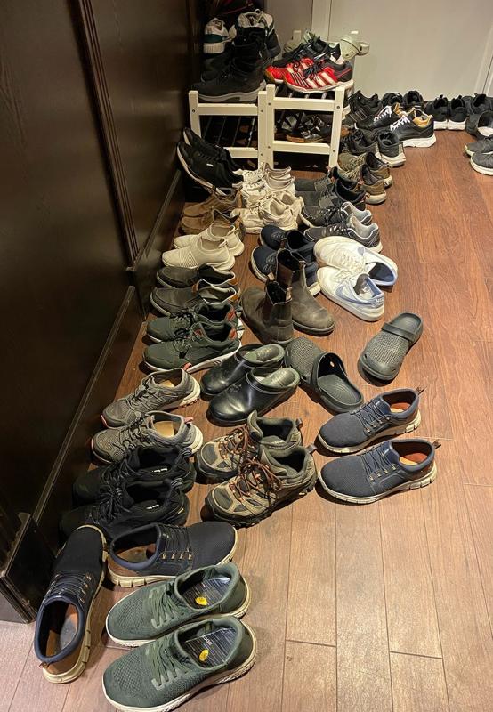 Många par skor, sandaler och sneakers ligger utspridda och staplade på hyllor och ett trägolv nära en mörk vägg.