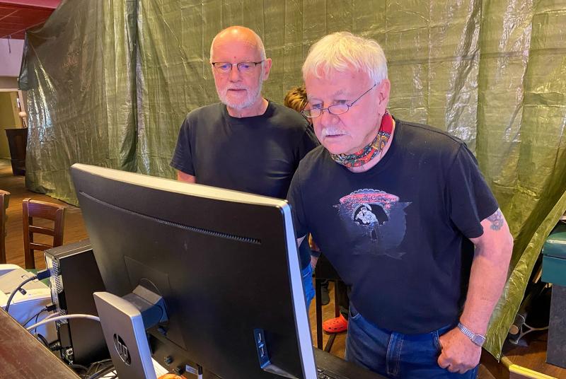 Två äldre män med glasögon och svarta t-shirts står tätt intill varandra och tittar på en datorskärm i ett rum med en presenning i bakgrunden.