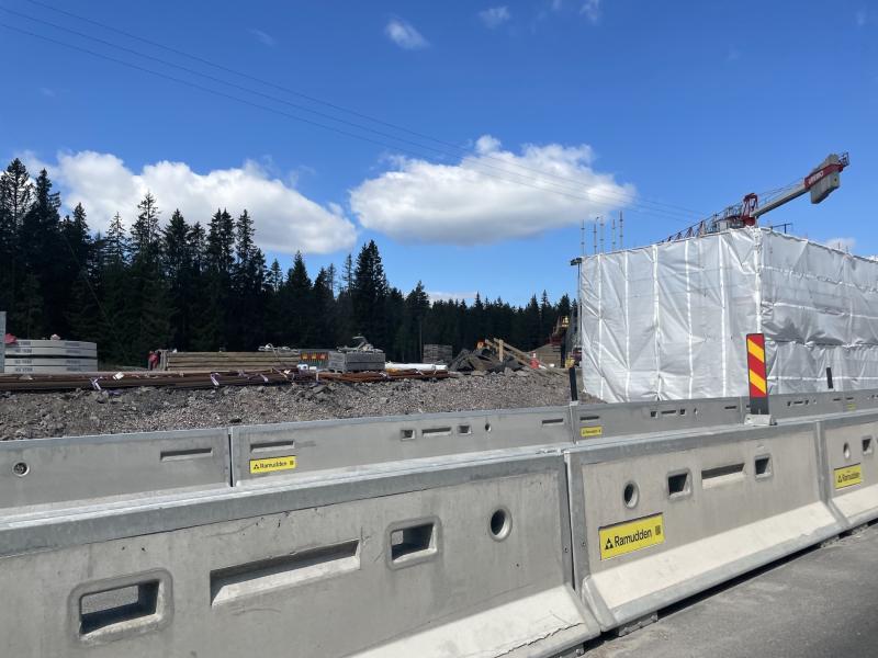 Byggarbetsplats med betongbarriärer i förgrunden, en inklämd struktur, byggmaterial och en kran; träd och en blå himmel med moln i bakgrunden.