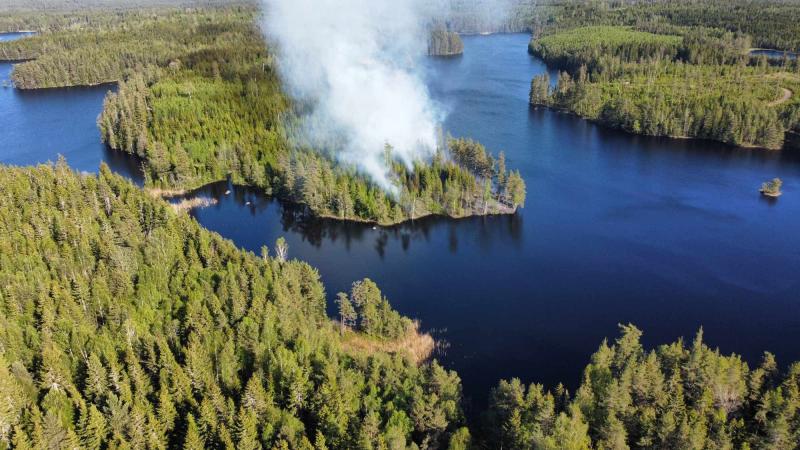 Flygfoto över en skogsbrand med rök som stiger från träd på en liten halvö omgiven av en sjö och tät skog.
