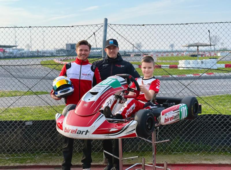 Tre personer poserar på en gokartbana; ett barn i racingutrustning sitter i en rödvit birelART gokart, flankerad av två vuxna, varav den ena håller i en hjälm.