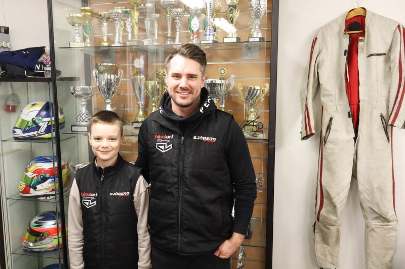 Två personer i racingvästar står framför en trofévitrin med hjälmar och en racingdräkt monterade på väggen.