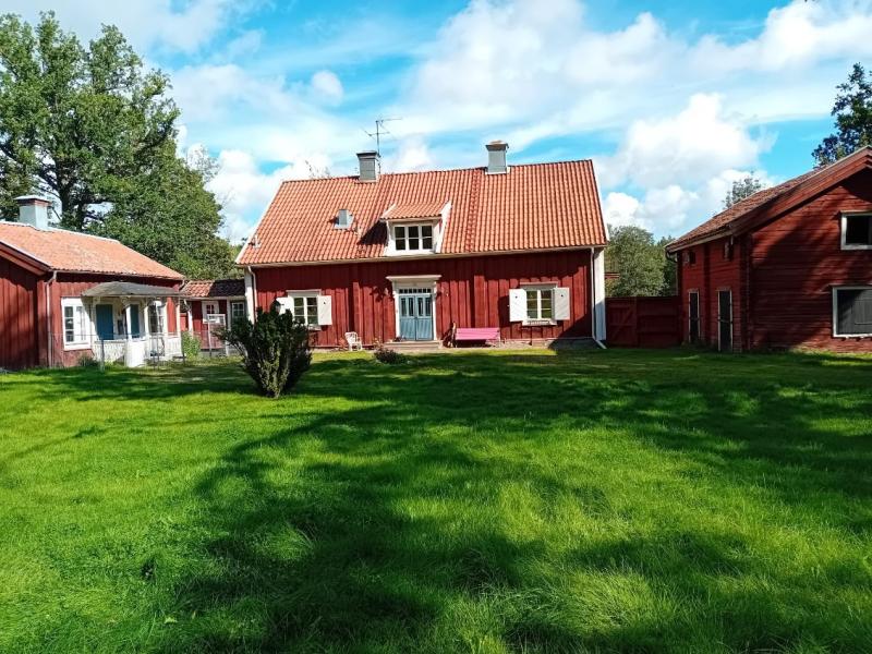 Ett traditionellt rött trähus med vita lister och tegeltak ligger på en grön gräsmatta under en delvis molnig himmel, omgivet av träd och uthus.