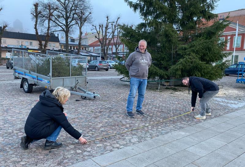 Två personer mäter ett avstånd på en kullerstensyta med ett måttband medan en annan person står i närheten; en husvagn med träd och byggnader syns i bakgrunden.