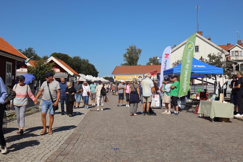 Folk promenerar och tittar på varorna vid stånden på en utomhusmarknad en solig dag. Flera tält och skyltar står uppställda längs en kullerstensgata med hus i bakgrunden.