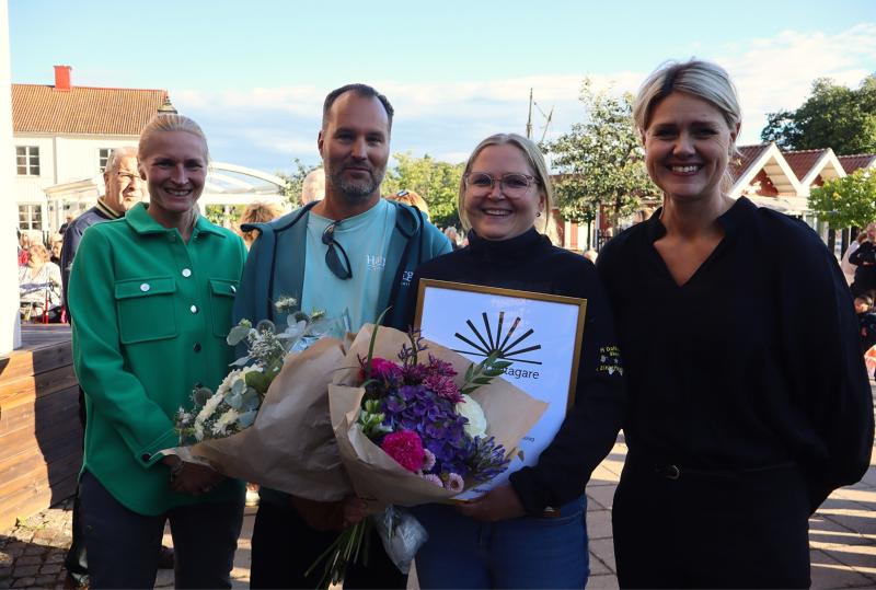 Fyra personer står utomhus; de två i mitten håller i blomsterbuketter och ett inramat certifikat. Alla ler. I bakgrunden syns hus och träd.
