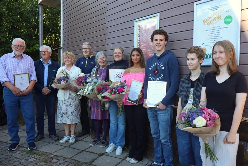 En grupp människor står på rad utomhus; några av dem håller i blomsterbuketter och diplom och poserar för ett foto framför en trävägg med affischer.