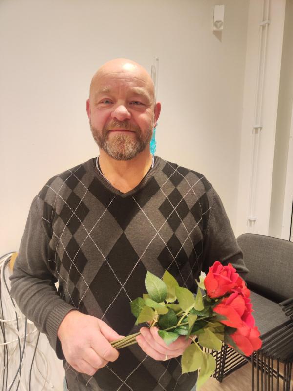 En man med skalligt huvud och skägg, klädd i en mörk argyle-tröja, står inomhus och håller i en bukett konstgjorda röda rosor.