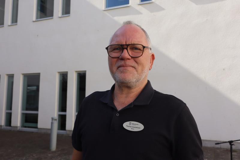 En äldre man med glasögon och en svart polotröja står utomhus framför en vit byggnad och tittar in i kameran.